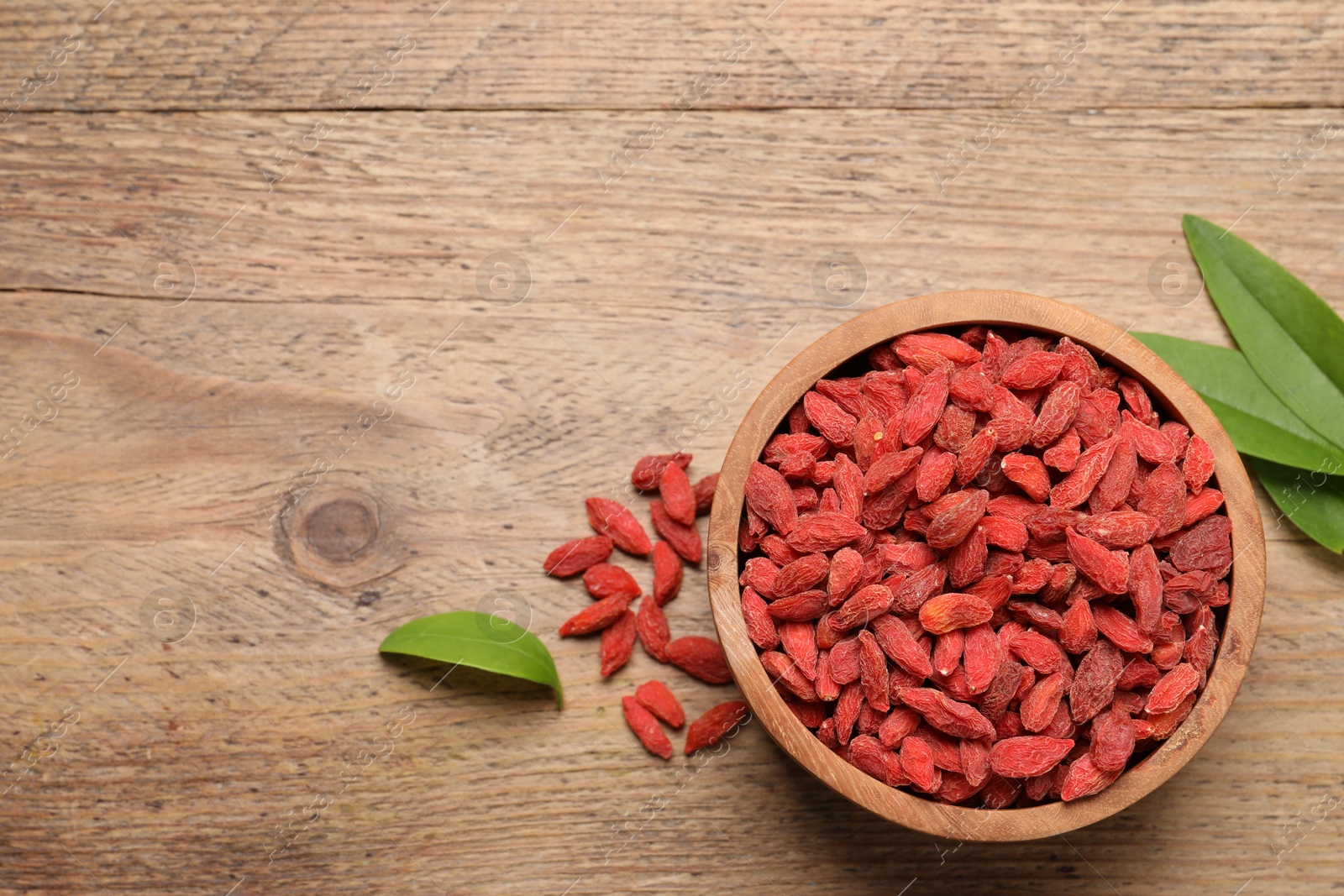 Photo of Dried goji berries, leaves and bowl on wooden table, flat lay. Space for text