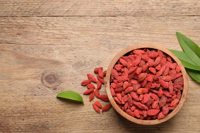 Dried goji berries, leaves and bowl on wooden table, flat lay. Space for text