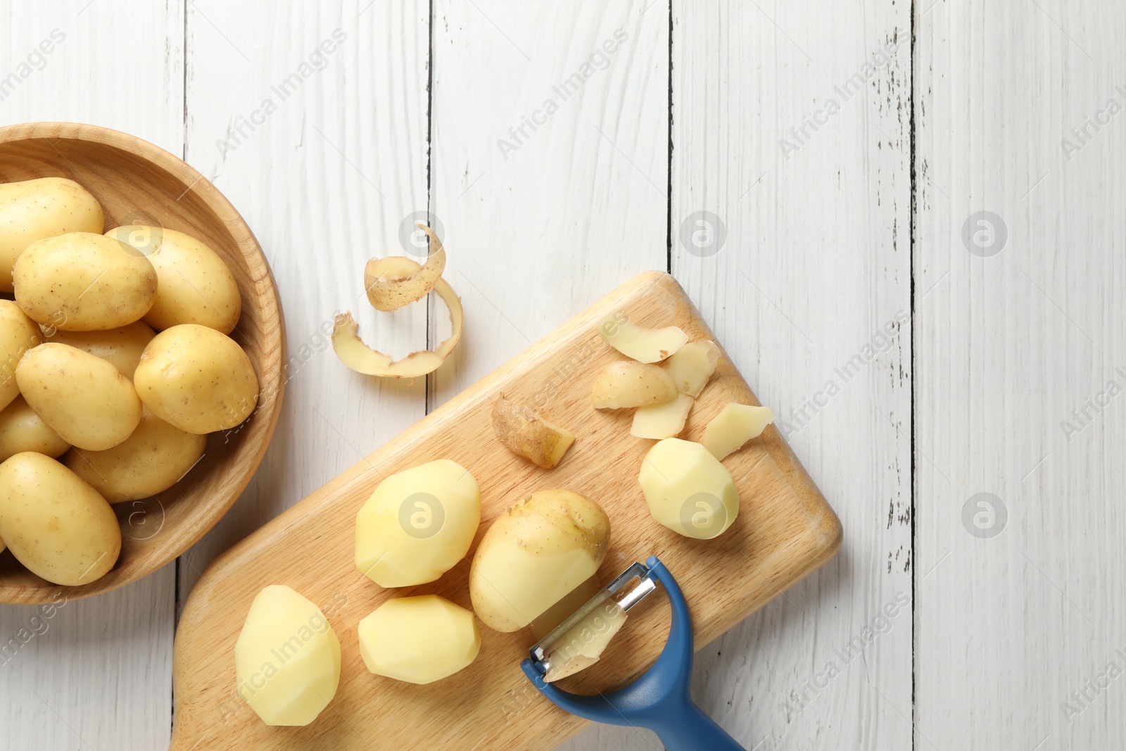 Photo of Fresh raw potatoes, peels and peeler on white wooden table, top view