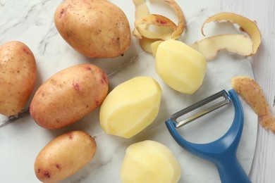 Fresh raw potatoes, peels and peeler on white wooden table, top view