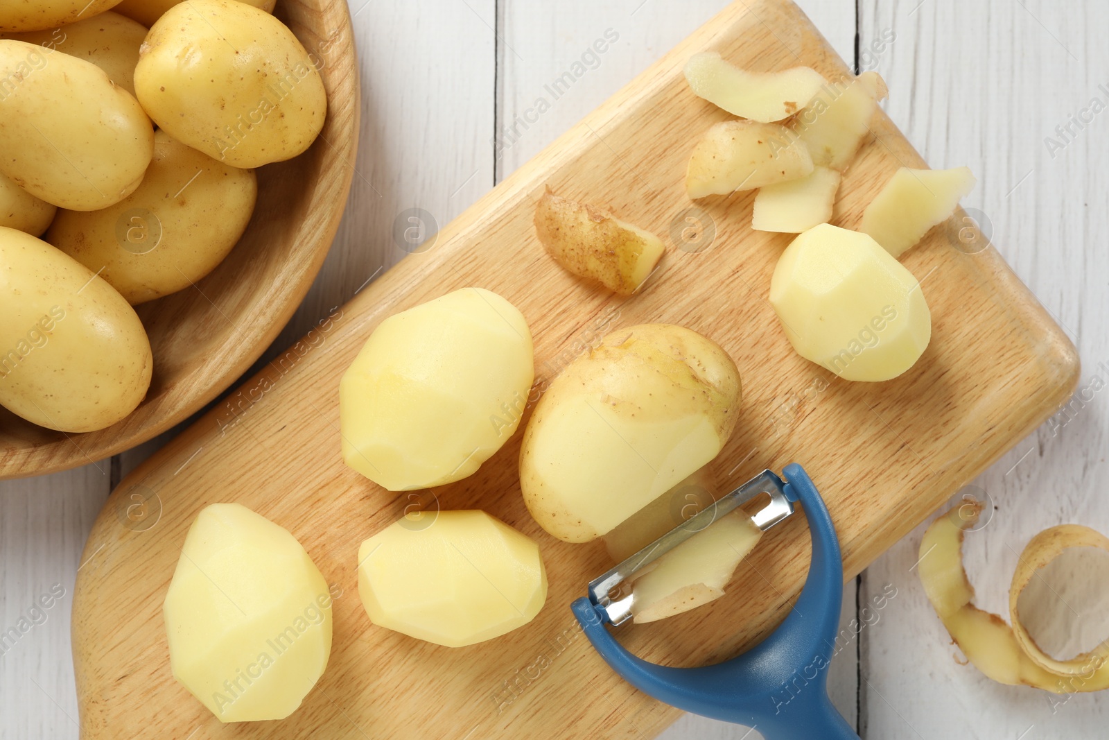 Photo of Fresh raw potatoes, peels and peeler on white wooden table, top view