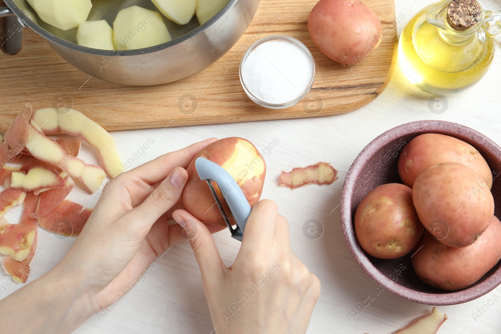 Photo of Woman peeling fresh potato with peeler at white wooden table, top view