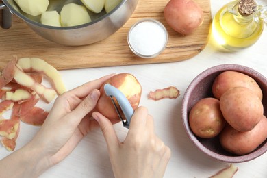 Photo of Woman peeling fresh potato with peeler at white wooden table, top view