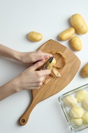 Woman peeling fresh potato with peeler at white table, top view