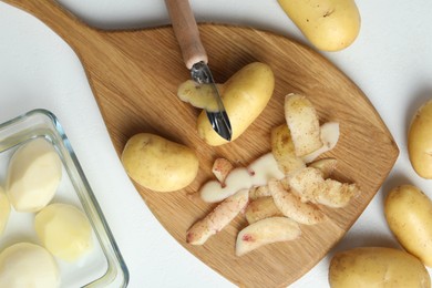 Photo of Fresh raw potatoes, peels and peeler on white table, top view