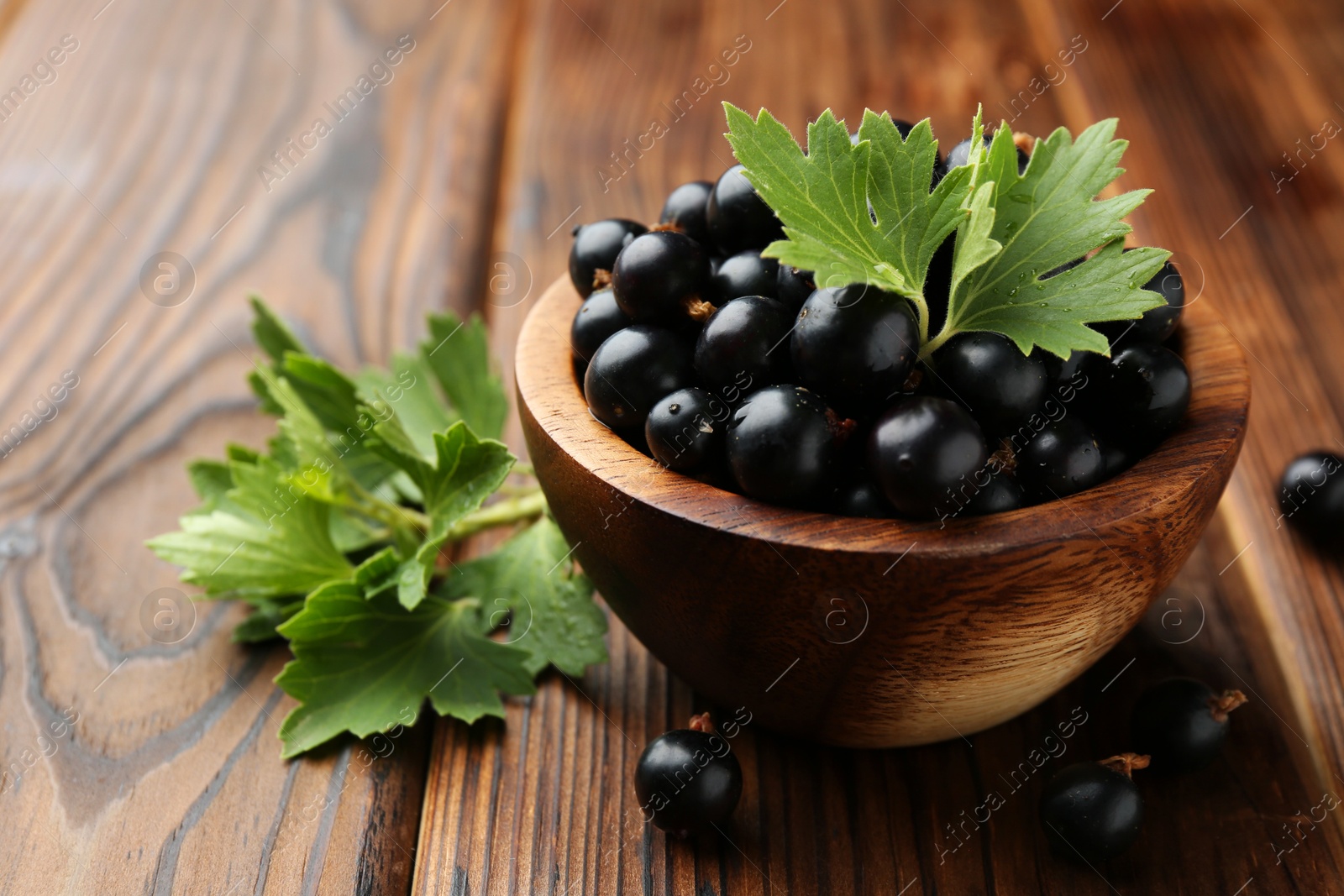 Photo of Ripe black currants and leaves in bowl on wooden table, closeup. Space for text