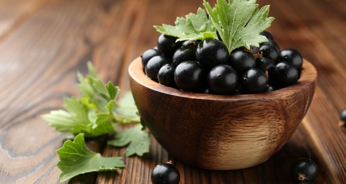 Photo of Ripe black currants and leaves in bowl on wooden table, closeup. Space for text