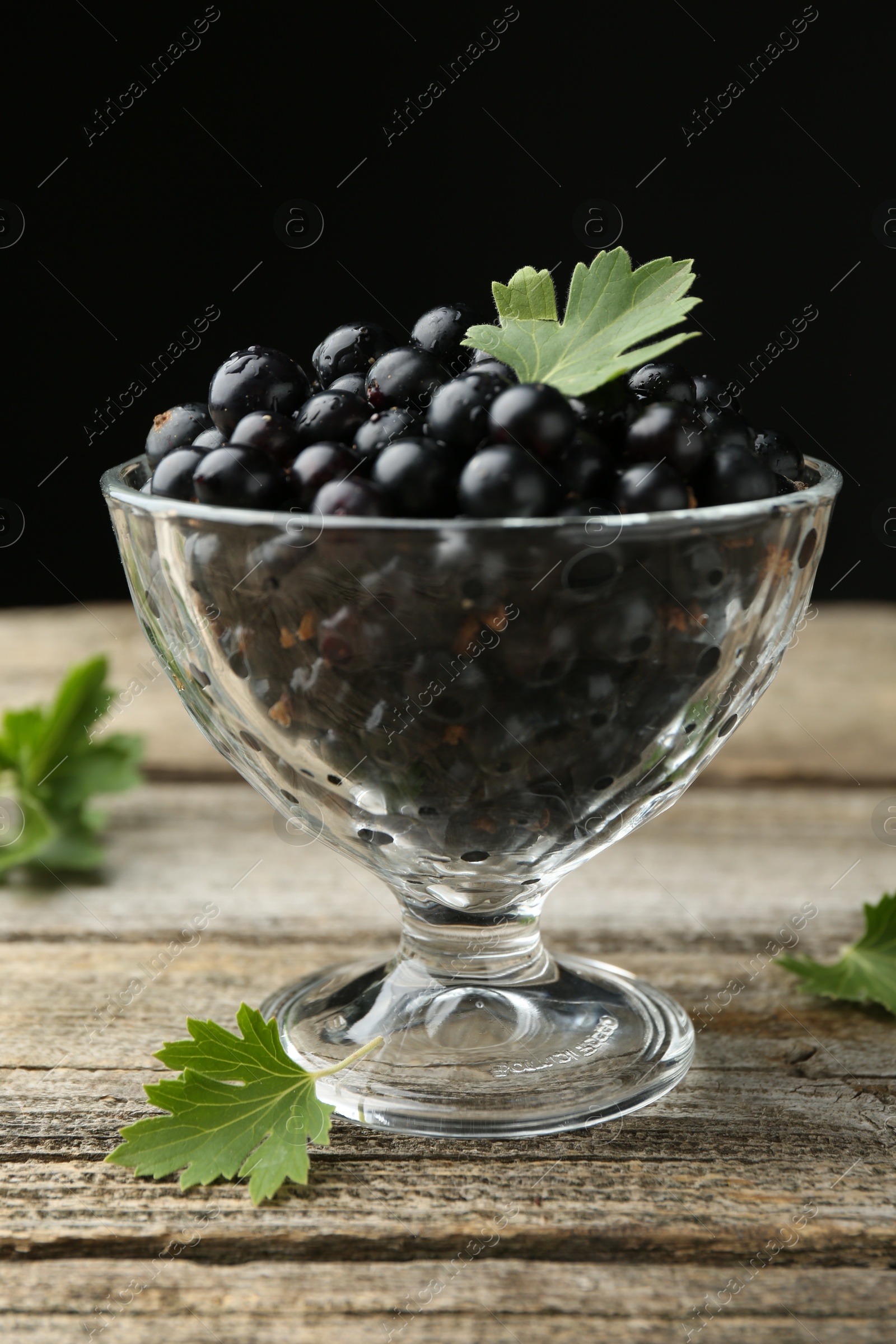 Photo of Ripe black currants and leaves in glass dessert bowl on wooden table