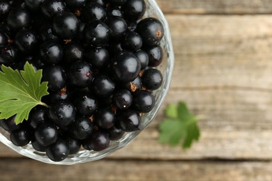 Photo of Ripe black currants and leaves in glass dessert bowl on wooden table, top view. Space for text