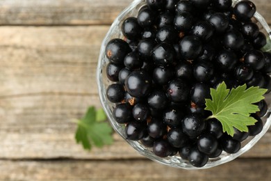 Photo of Ripe black currants and leaves in glass dessert bowl on wooden table, top view. Space for text