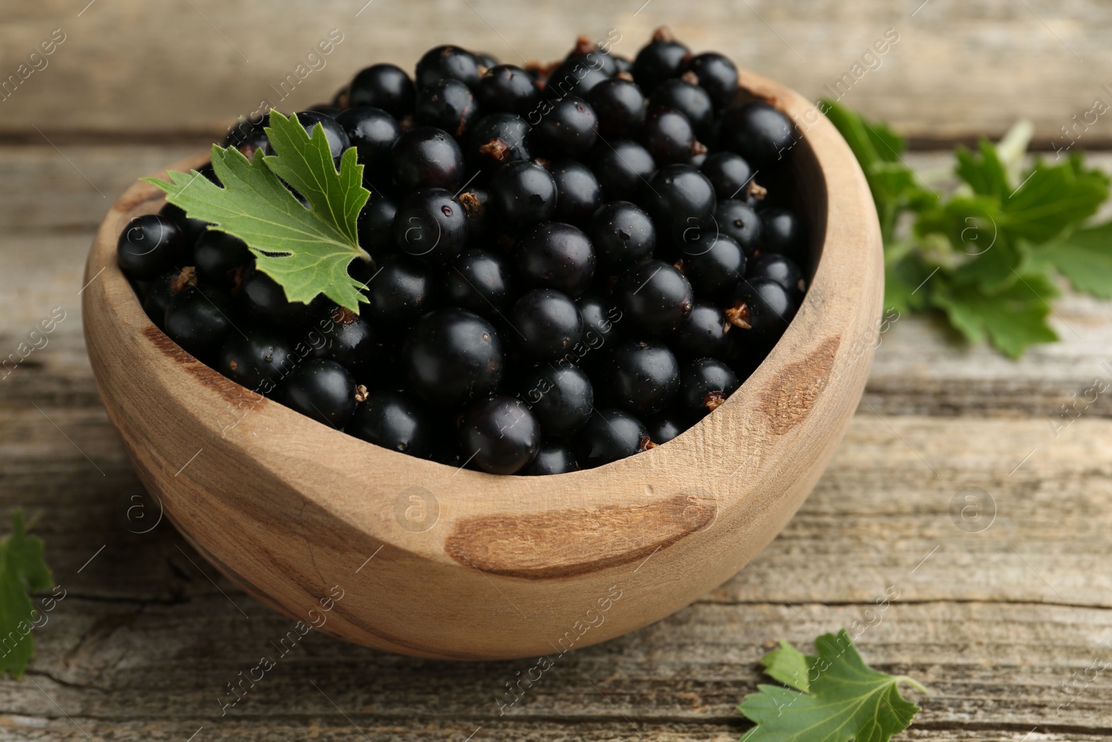 Photo of Ripe black currants and leaves in bowl on wooden table, closeup