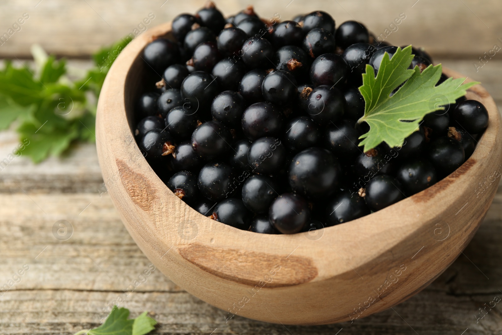 Photo of Ripe black currants and leaves in bowl on wooden table, closeup