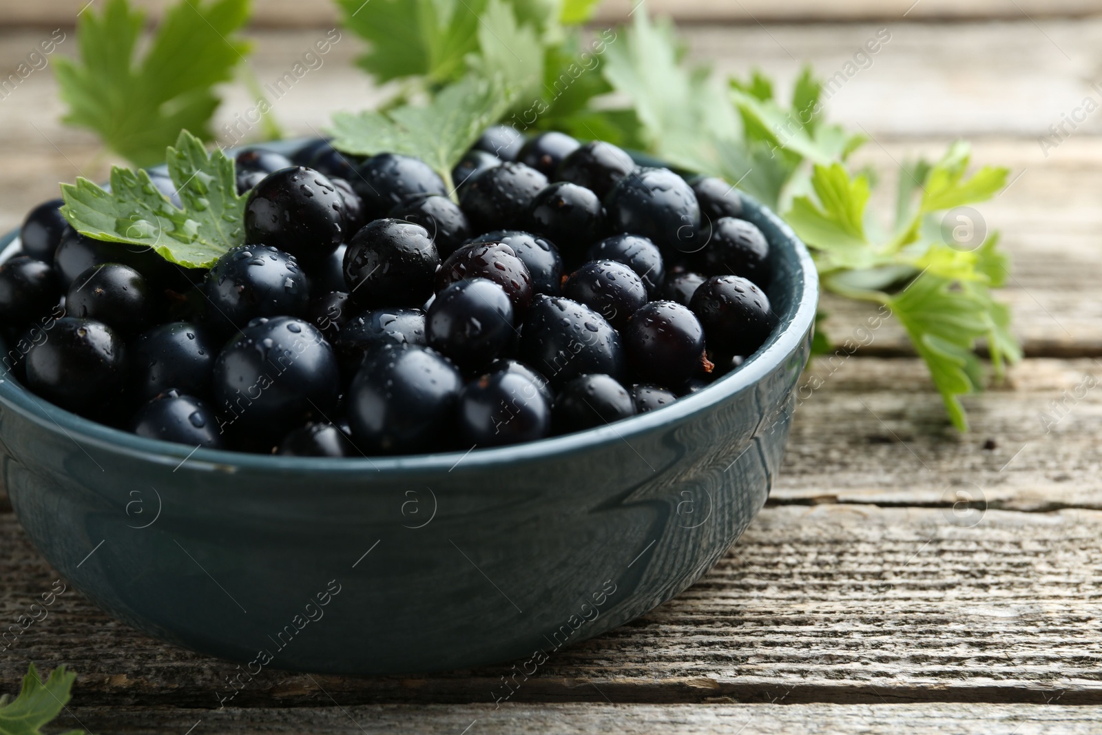 Photo of Ripe blackcurrants in bowl and green leaves on wooden table, closeup