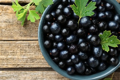 Photo of Ripe blackcurrants in bowl and green leaves on wooden table, top view