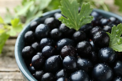 Ripe blackcurrants in bowl and green leaves on wooden table, closeup