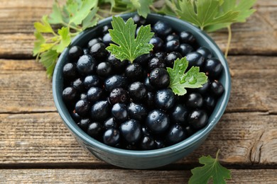 Photo of Ripe blackcurrants in bowl and green leaves on wooden table