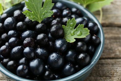 Photo of Ripe blackcurrants in bowl and green leaves on wooden table, closeup