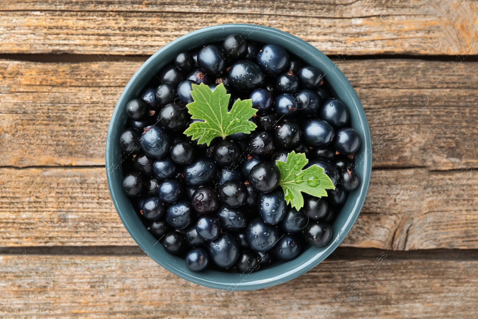 Photo of Ripe blackcurrants in bowl and green leaves on wooden table, top view