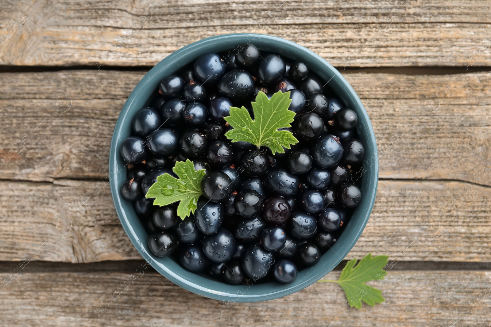 Photo of Ripe blackcurrants in bowl and green leaves on wooden table, top view