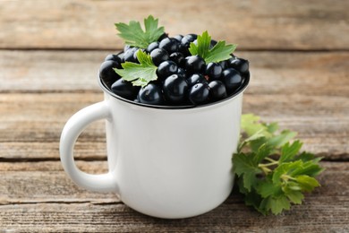 Photo of Ripe blackcurrants in mug and green leaves on wooden table, closeup