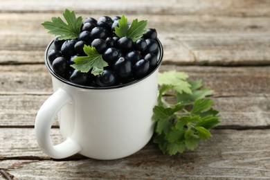 Photo of Ripe blackcurrants in mug and green leaves on wooden table, closeup