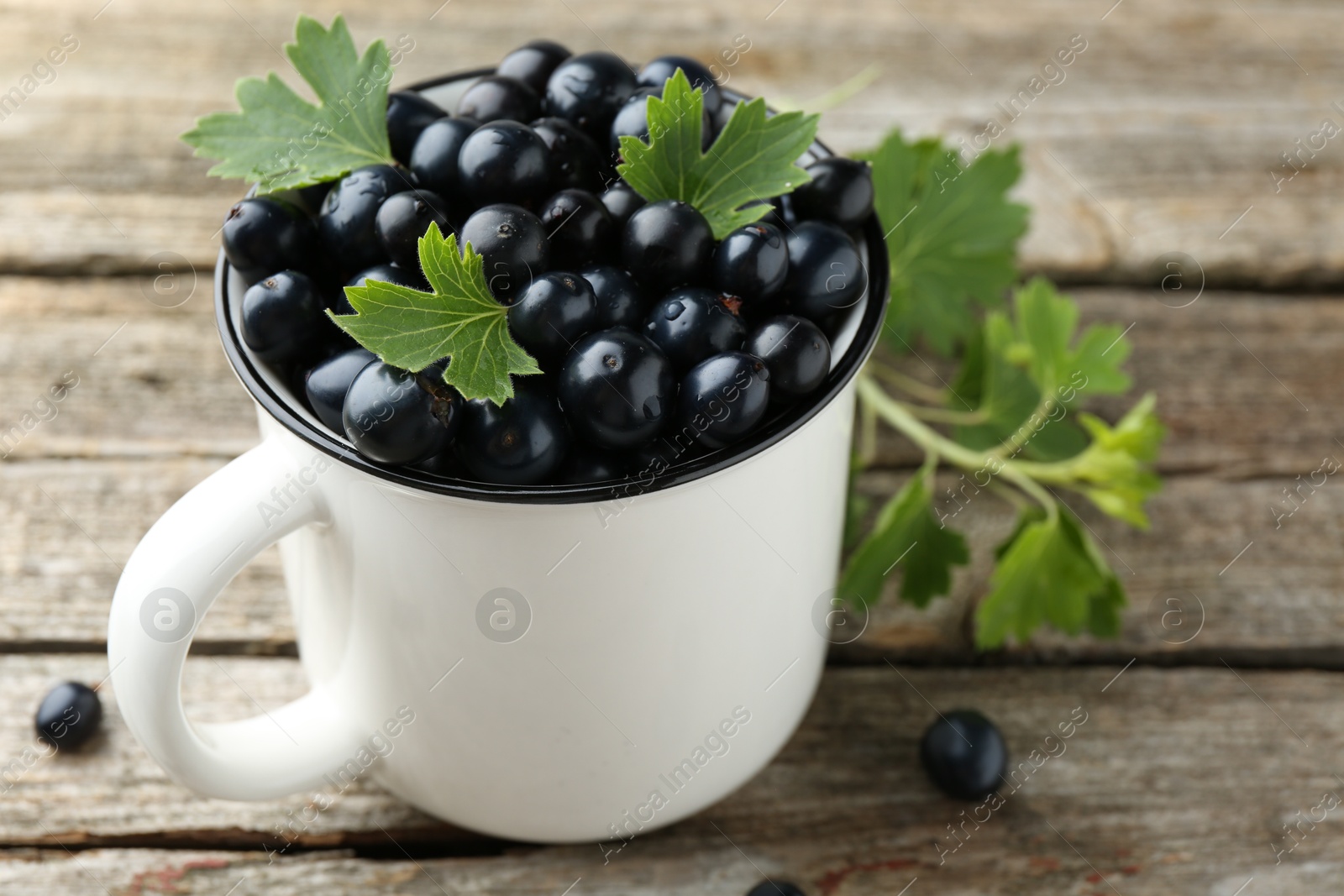 Photo of Ripe blackcurrants in mug and green leaves on wooden table, closeup