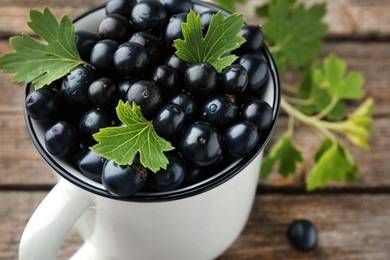 Ripe blackcurrants in mug and green leaves on wooden table, closeup