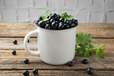 Photo of Ripe blackcurrants in mug and green leaves on wooden table