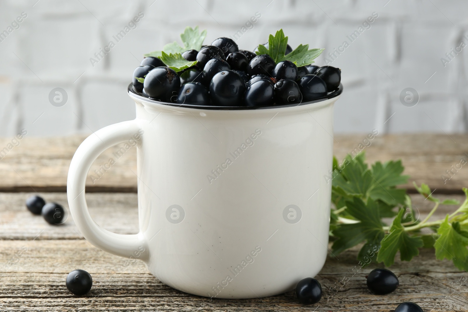 Photo of Ripe blackcurrants in mug and green leaves on wooden table, closeup