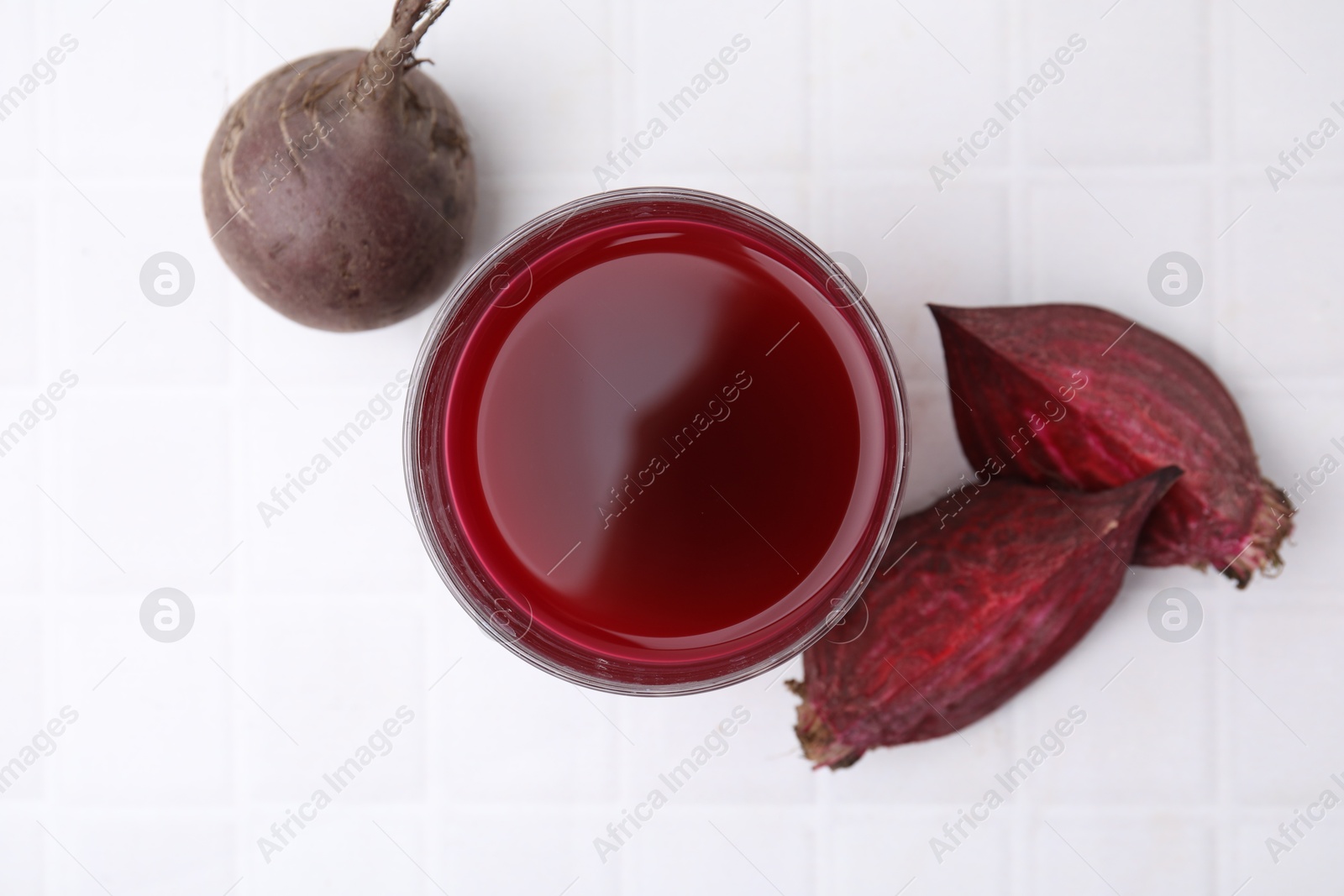 Photo of Fresh beet juice in glass and ripe vegetables on white tiled table, flat lay