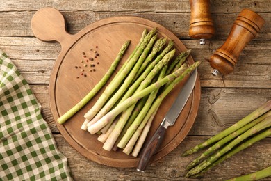 Photo of Board with fresh green asparagus stems, spices and knife on wooden table, flat lay