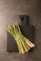 Photo of Board with bunch of fresh green asparagus stems on grey textured table, top view