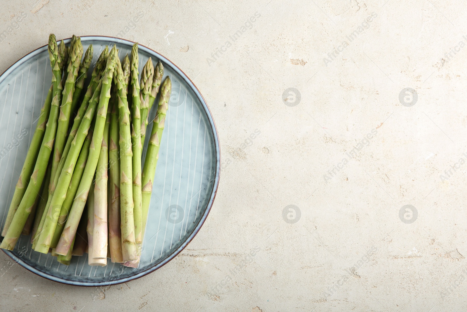 Photo of Plate with fresh green asparagus stems on light textured table, top view. Space for text