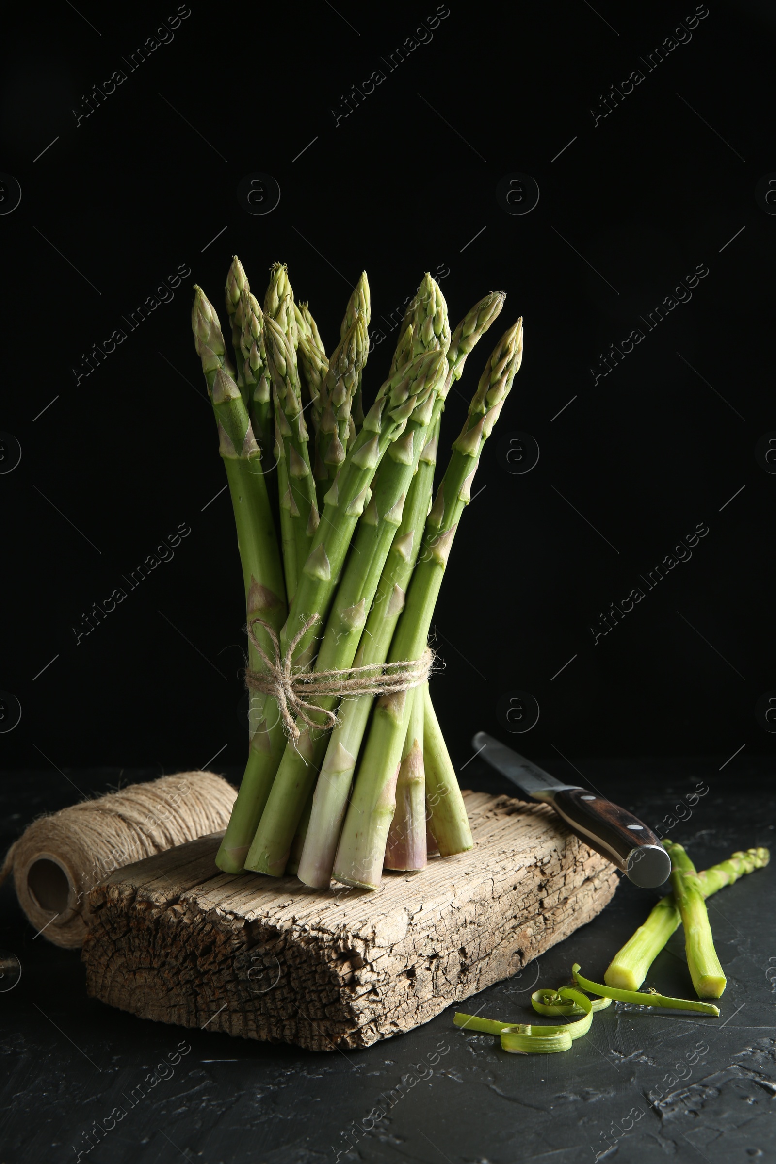 Photo of Fresh green asparagus stems, thread and knife on gray textured table against black background