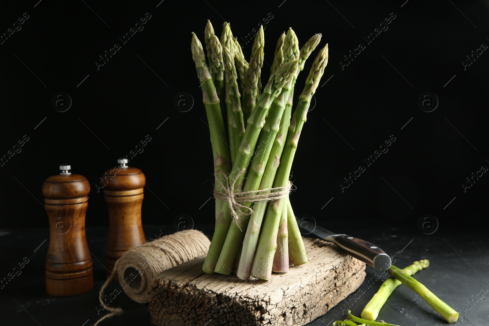 Photo of Fresh green asparagus stems, spices, thread and knife on gray textured table against black background