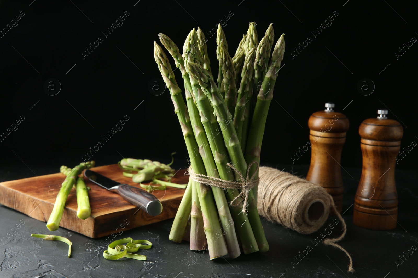 Photo of Fresh green asparagus stems, spices, thread and knife on gray textured table against black background