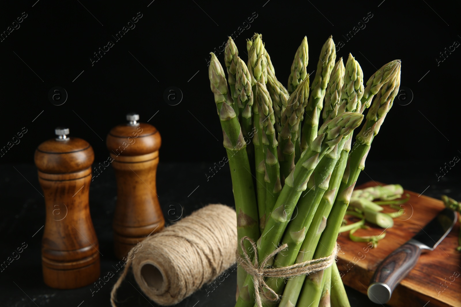 Photo of Fresh green asparagus stems, spices, thread and knife on table against black background