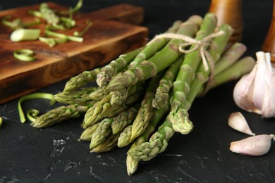 Photo of Fresh green asparagus stems and garlic on black textured table, closeup