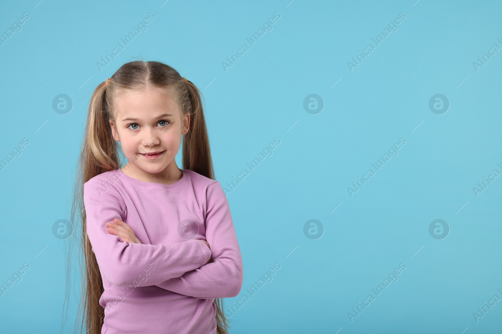 Photo of Portrait of cute little girl with crossed arms on light blue background, space for text