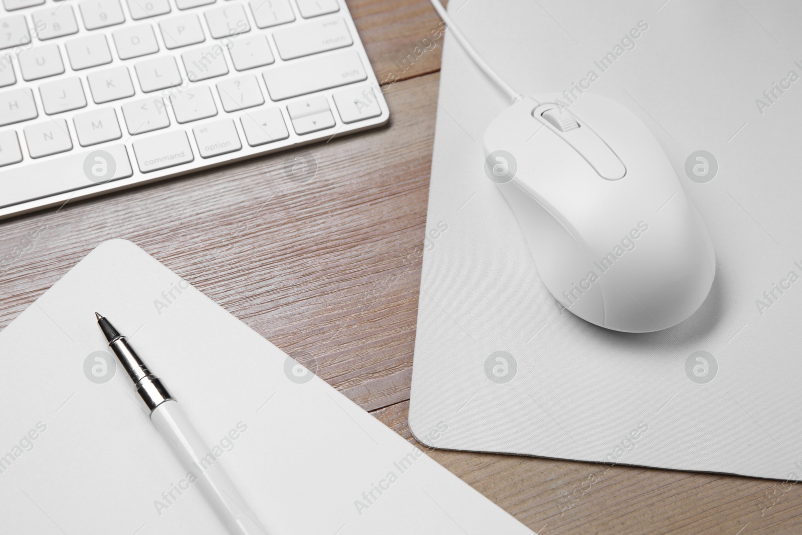 Photo of Wired mouse with mousepad, notebook, pen and computer keyboard on wooden table, above view