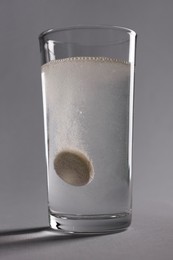 Effervescent pill dissolving in glass of water on grey background