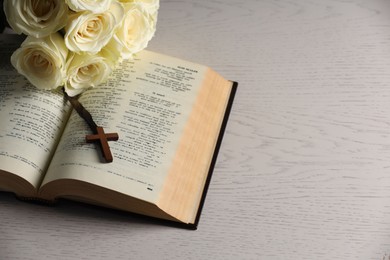 Photo of Bible, cross and roses on light wooden table, space for text. Religion of Christianity