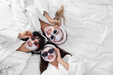 Photo of Happy friends with facial masks wearing sunglasses on bed, top view and space for text. Spa party