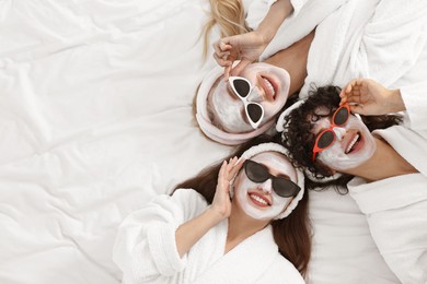 Photo of Happy friends with facial masks wearing sunglasses on bed, top view and space for text. Spa party