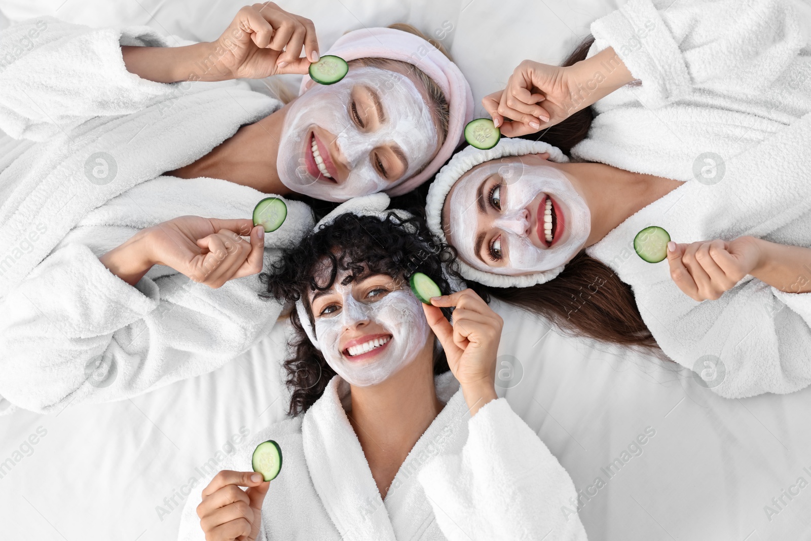 Photo of Happy friends with facial masks and cucumber slices on bed, top view. Spa party