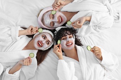 Happy friends with facial masks and cucumber slices on bed, top view. Spa party