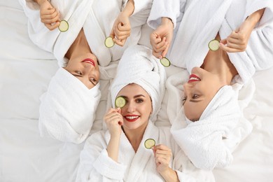 Happy friends in bathrobes with cucumber slices on bed, top view. Spa party