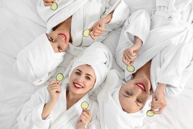 Photo of Happy friends in bathrobes with cucumber slices on bed, top view. Spa party