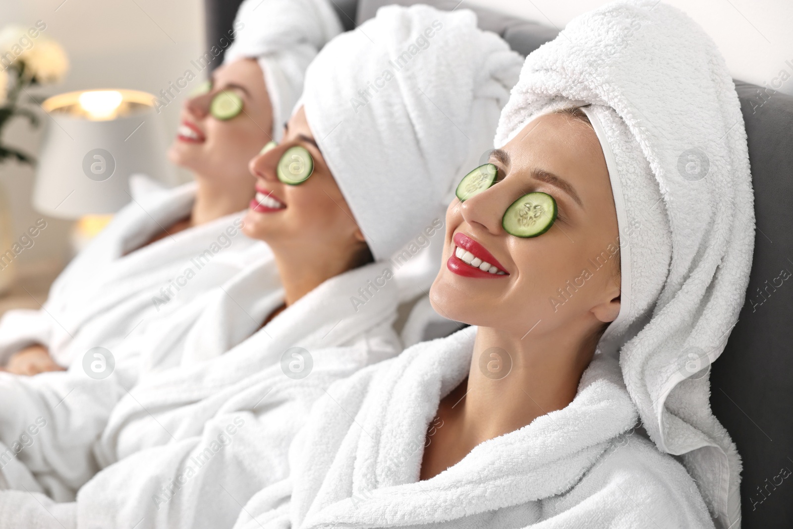 Photo of Happy friends in bathrobes with cucumber slices on bed, selective focus. Spa party