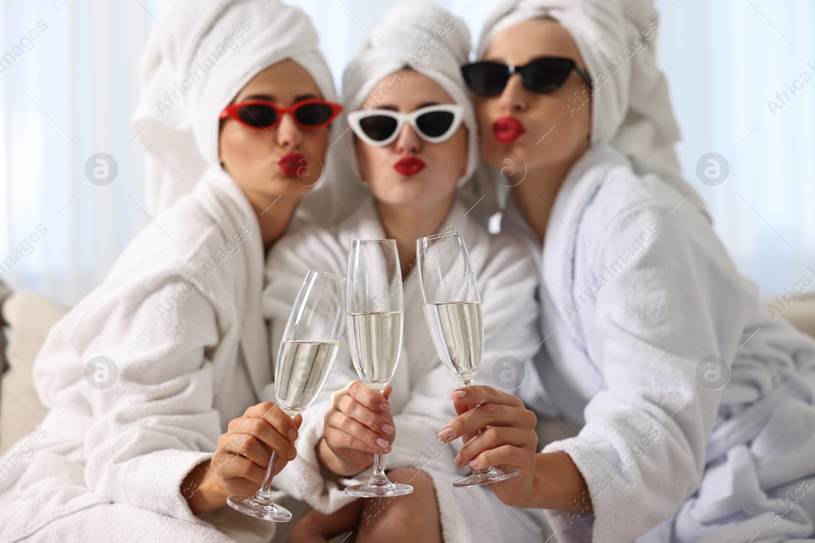 Photo of Happy friends in bathrobes with sunglasses and glasses of sparkling wine indoors, selective focus. Spa party